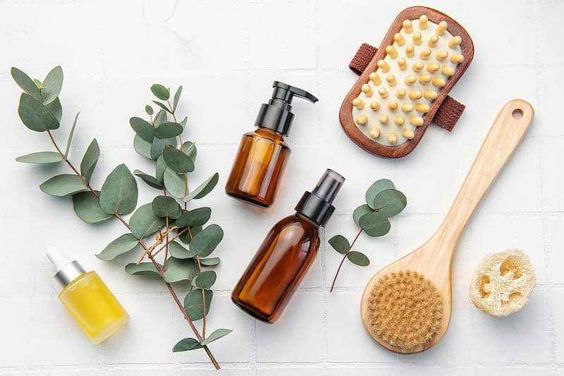 Natural wellness products including amber glass bottles, a wooden body brush, a massage brush, a small dropper bottle, eucalyptus branches, and a loofah arranged on a white background