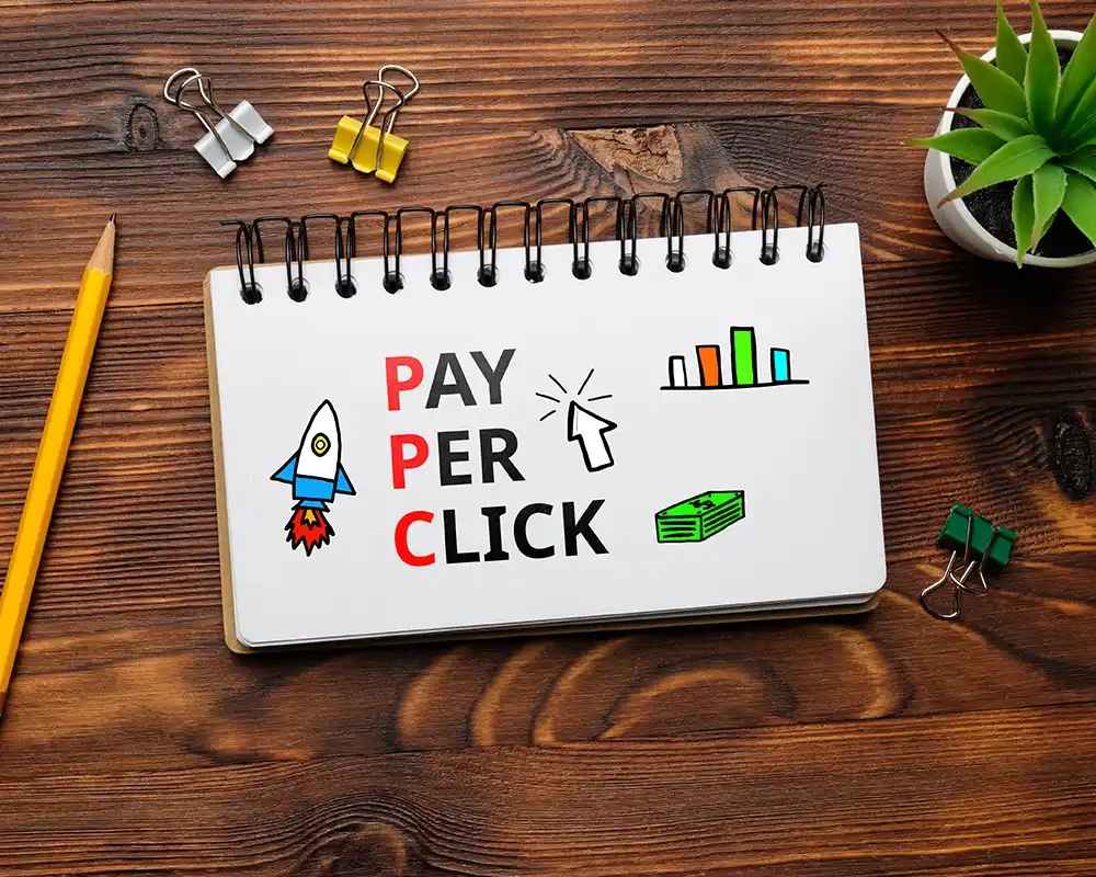 Notebook with "Pay Per Click" text, rocket icon, mouse cursor, bar chart, and money graphic, representing PPC advertising strategies.